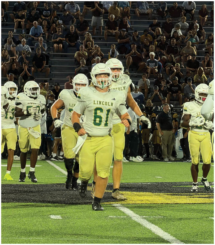 A group of football players in white and yellow uniforms with helmets are walking on a field with spectators in the background. Description generated by AI