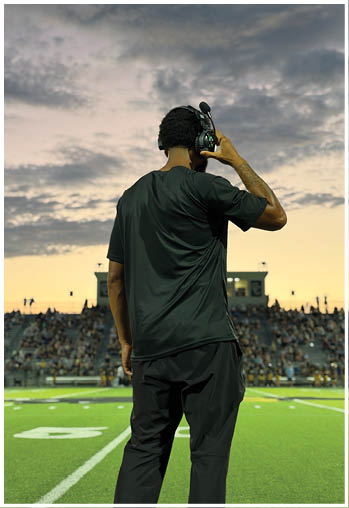 A person wearing a headset and dark clothing stands on a football field facing a crowd in the stadium during sunset. Description generated by AI