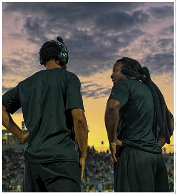 An adult wearing a dark head covering and shirt is crouched in a ready position on a sports field with another person in green sports gear blurred in the background. Description generated by AI