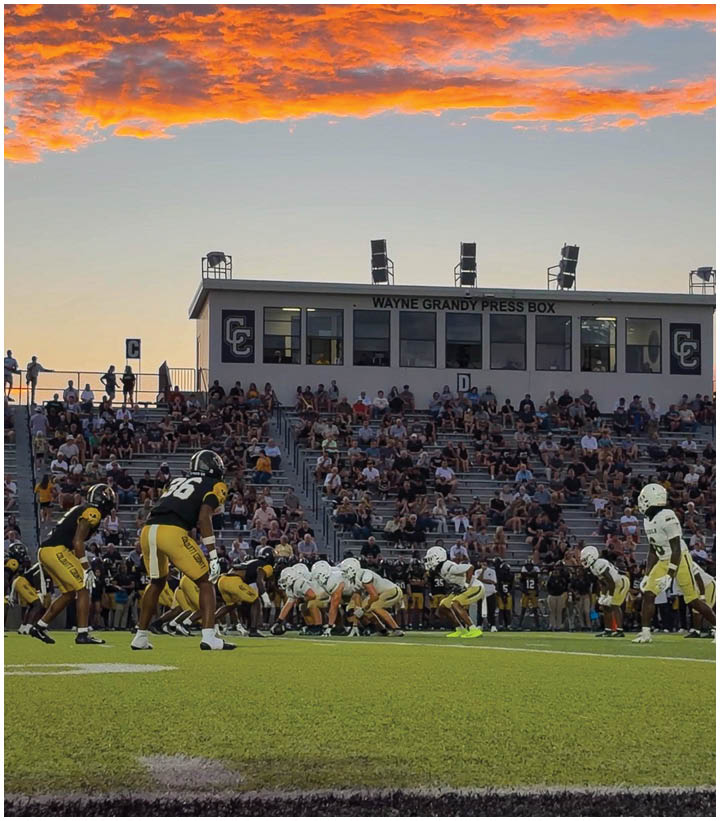 A football game is taking place at sunset with players in yellow and white uniforms lined up on the field and a crowd watching from the bleachers under a vibrant orange and purple sky. Description generated by AI