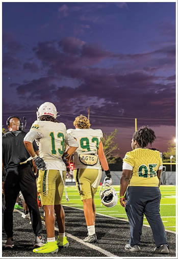 Three people in football uniforms stand on the sidelines of a field under a darkening sky with clouds. Description generated by AI