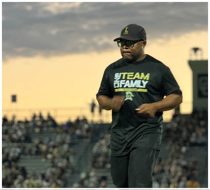A person wearing a black cap and a black t-shirt with the words “TEAM FAMILY" is jogging or running outdoors in front of a blurred crowd during sunset. Description generated by AI