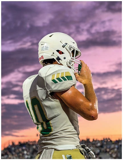 A person wearing a white football uniform and helmet is standing on a field with a sunset sky in the background. Description generated by AI