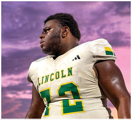 A person wearing a white football jersey with the word Lincoln and the number 72 stands against a purple and pink cloudy sky. Description generated by AI