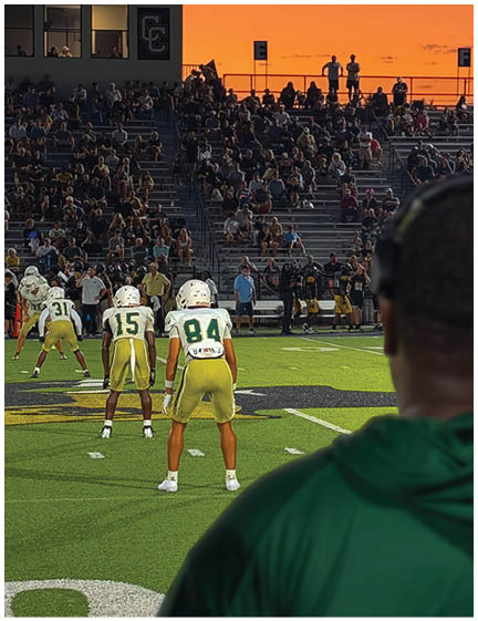 A person wearing a green hoodie and headphones watches a group of people in football uniforms preparing on a field with a crowd in the stands during a sunset. Description generated by AI