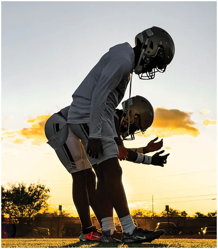 Two people wearing football helmets and athletic clothing are crouched in a ready position on a field at sunset. Description generated by AI