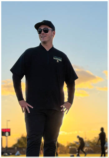 A person wearing sunglasses, a black cap, and a black shirt with “Tiblan Nation" printed on it is standing outdoors at sunset with two other people walking in the background. Description generated by AI