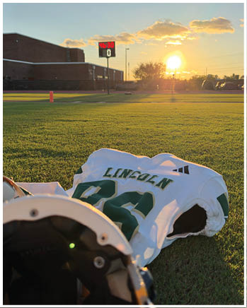 A football jersey with the name Lincoln is laid on the grass of a sports field at sunset with a scoreboard and buildings in the background. Description generated by AI