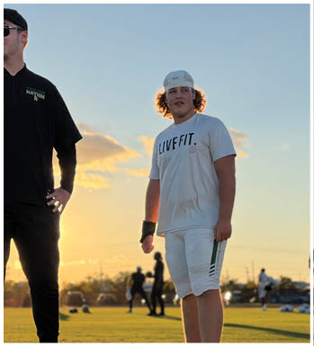 Two adults stand on a grassy field at sunset, one wearing a black outfit with sunglasses and a cap, and the other wearing a white athletic outfit with a backward cap. Description generated by AI