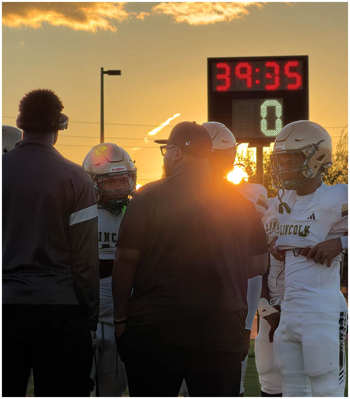 People wearing football helmets and gear are gathered near a scoreboard at sunset with a partly cloudy sky. Description generated by AI