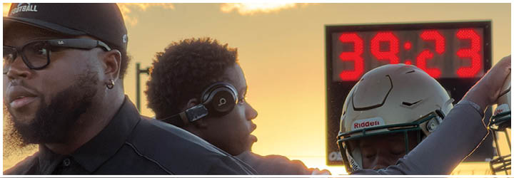 Two people wearing Lincoln football gear are interacting, with one person adjusting the other's helmet in a setting illuminated by warm sunlight. Description generated by AI