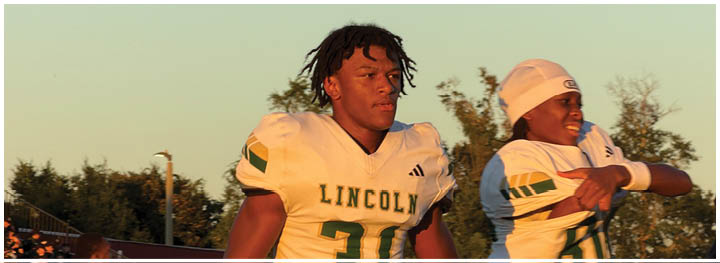 Two adults wearing football uniforms with “LINCOLN" on the jerseys are on a field with bleachers and trees in the background during sunset. Description generated by AI