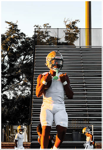 A person wearing a football helmet and white athletic gear is standing on a field with other similarly dressed people and bleachers in the background. Description generated by AI