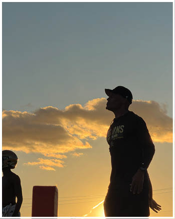 Two people stand outdoors at sunset with one wearing a cap and the other wearing a helmet against a sky with scattered clouds. Description generated by AI