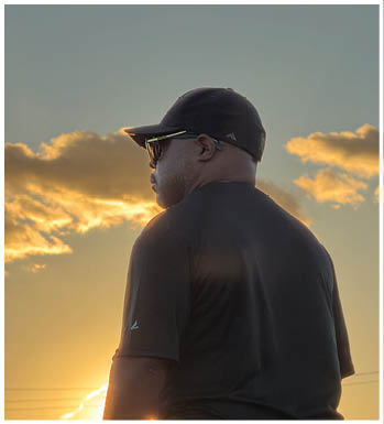 A person wearing a black cap, sunglasses, and a black shirt is standing outdoors with a sunset and clouds in the background. Description generated by AI