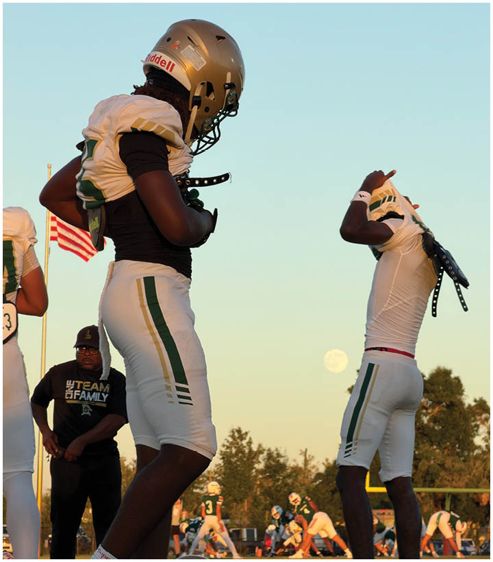 Two people in football uniforms and helmets stand on a field at sunset with an American flag and a person in casual clothing in the background. Description generated by AI