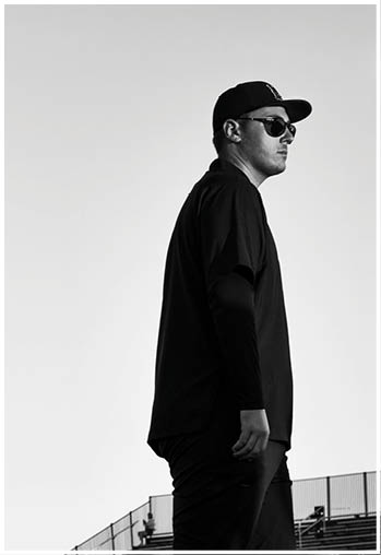 A person wearing sunglasses, a cap, and dark clothing is standing in profile against a clear sky. Description generated by AI