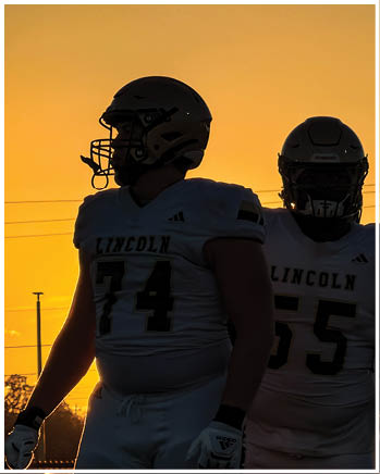 Two people wearing football uniforms with helmets stand against a sunset background. Description generated by AI