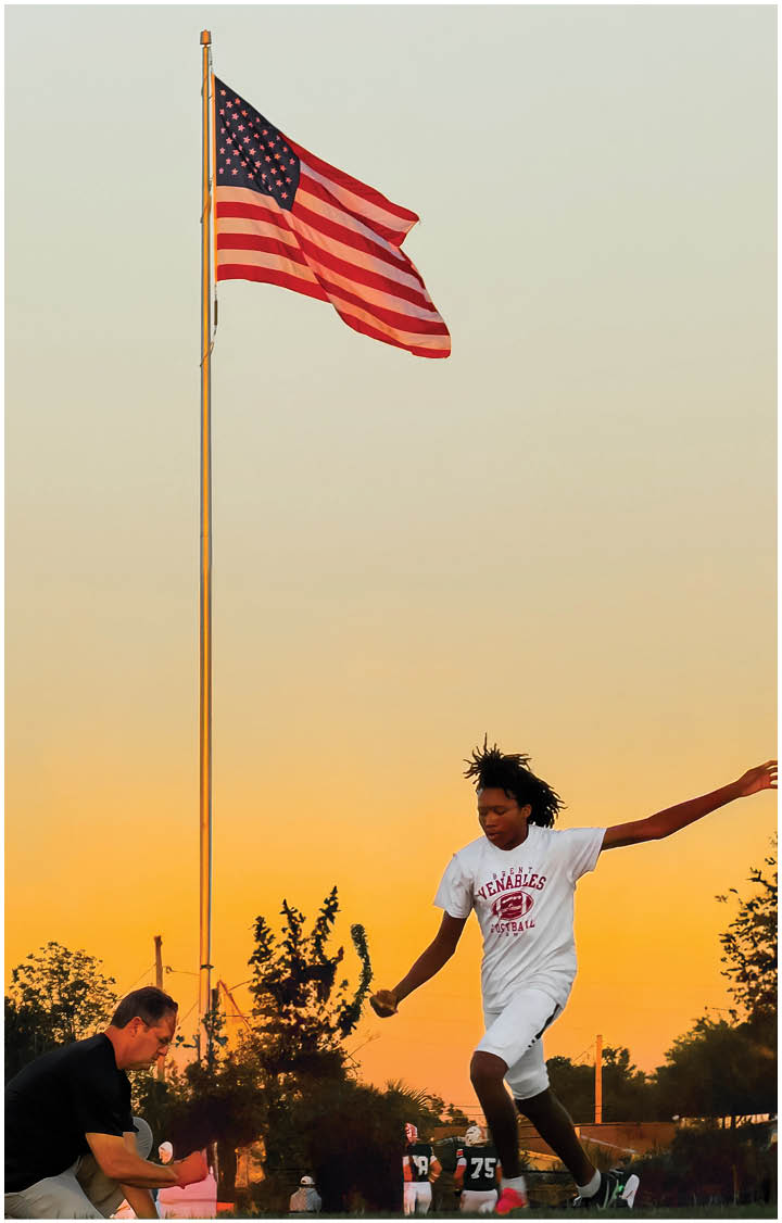 A person with dreadlocks wearing a white sports t-shirt is walking or running outdoors near a tall flagpole with an American flag against a sunset sky. Description generated by AI