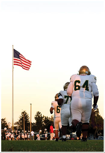 Several people in football uniforms stand near a tall flagpole with an American flag waving against a clear sky. Description generated by AI
