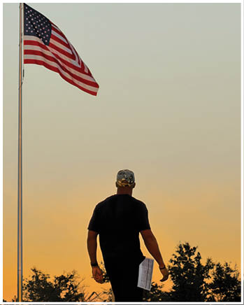 A person wearing a camouflage hat and black shirt stands outdoors near a flagpole with an American flag against a clear sky at sunset. Description generated by AI