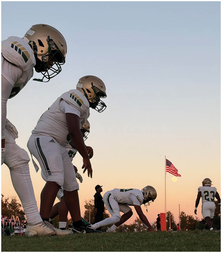 Several football players in helmets and uniforms are lined up on a grassy field at sunset with an American flag and a person standing in the background. Description generated by AI