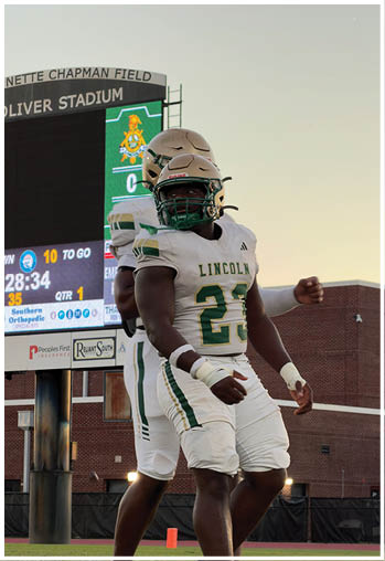 Two people wearing football uniforms and helmets are standing on a field near a scoreboard and stadium seating during a game. Description generated by AI