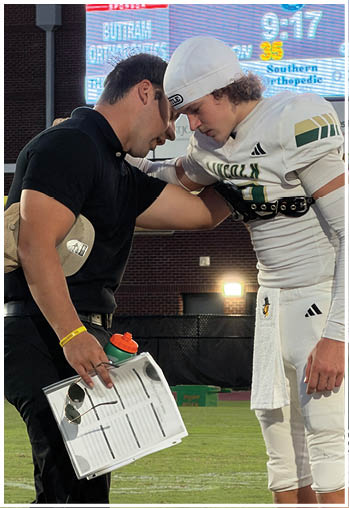 An adult wearing a black shirt is leaning in and speaking closely to a person in a white football uniform and helmet on a sports field. Description generated by AI