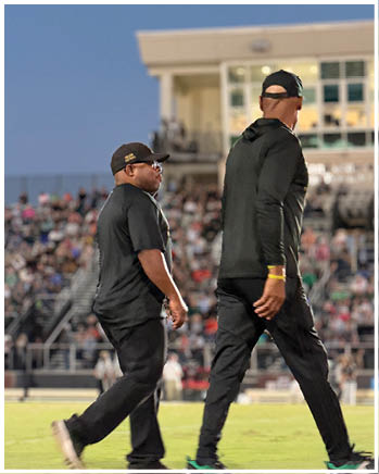 Two adults wearing black clothing and caps are walking side by side in a stadium with a crowd in the background. Description generated by AI