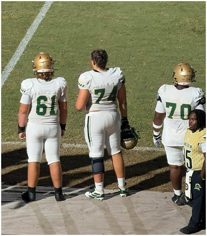 Four people stand on the sidelines of a football field, three wearing white football uniforms with helmets and one in casual clothing. Description generated by AI