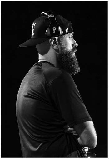 A person with a beard wearing a backward cap and headphones is standing with arms crossed against a dark background. Description generated by AI