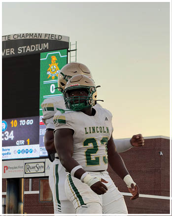 Two people wearing white and green football uniforms and gold helmets stand on a field near a scoreboard at sunset. Description generated by AI