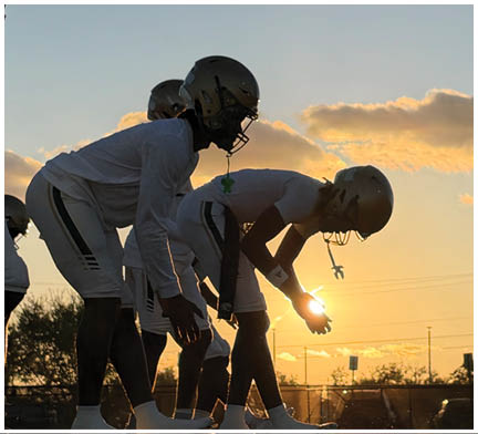 People wearing football helmets and uniforms are lined up on a field at sunset preparing for a play. Description generated by AI