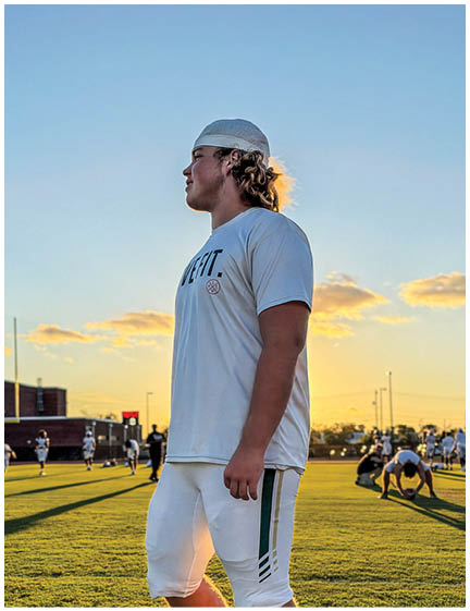 A person wearing a white sports outfit and cap stands on a grassy field at sunset while others practice or play sports in the background. Description generated by AI