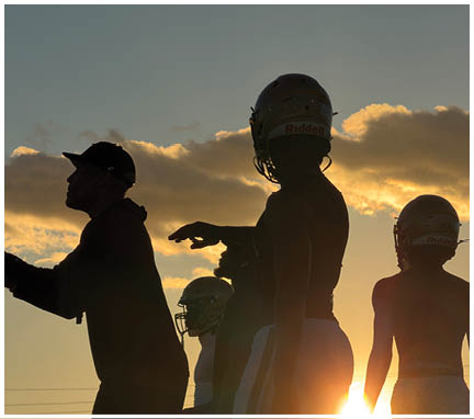 Three people are silhouetted against a sunset sky, with one wearing a football helmet and another wearing a cap. Description generated by AI