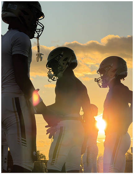 Three people wearing football helmets and uniforms stand together on a field with the sun setting behind them. Description generated by AI