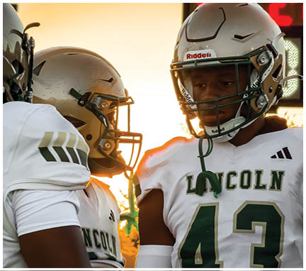 Two people wearing football helmets and white jerseys with “LINCOLN" and the number 43 are standing close together during sunset. Description generated by AI
