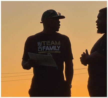 Two people stand facing each other outdoors at sunset, with one holding a piece of paper and wearing a cap and a shirt that says “TEAM FAMILY." Description generated by AI