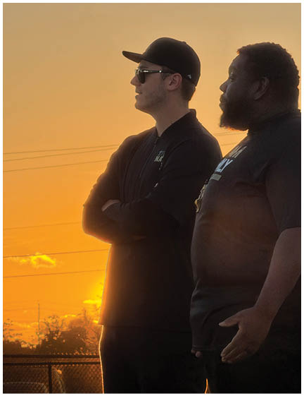 Two adults wearing dark clothing stand side by side outdoors with a sunset sky in the background. Description generated by AI