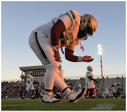 A person in a football uniform and helmet is bending forward on a field with other players and spectators in the background during sunset. Description generated by AI