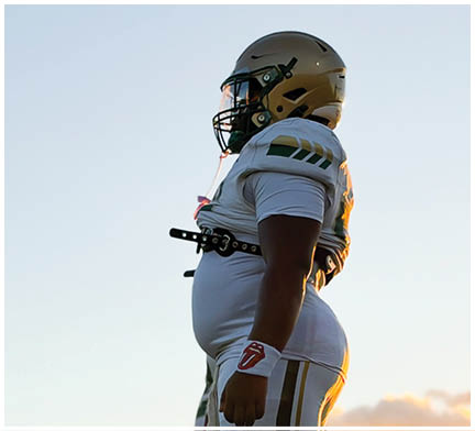 An adult wearing a white football uniform with the number 51 and the word Lincoln walks on a field with empty bleachers and a press box in the background. Description generated by AI