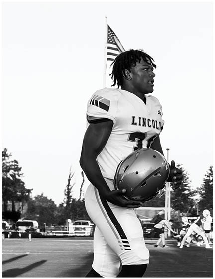 A person wearing a Lincoln football uniform is holding a helmet and standing on a field with an American flag in the background. Description generated by AI