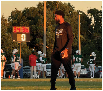 A person in a black long-sleeve shirt holding a football stands on a field with several people in football uniforms and a scoreboard showing time and score in the background. Description generated by AI