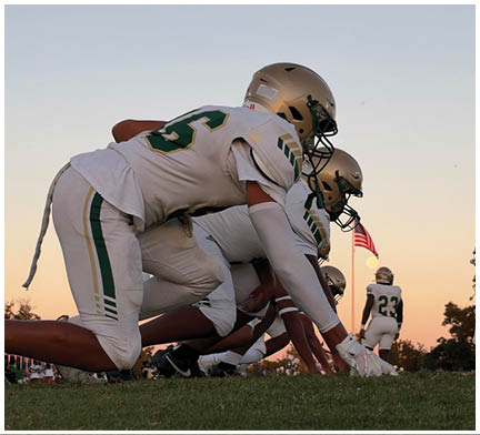 Two people in football uniforms and helmets are crouched in a ready position on a field with an American flag and a clear sky in the background. Description generated by AI