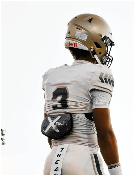 A person wearing a white football uniform with the number 3 and a gold helmet stands on a field with a black protective pad strapped to their lower back. Description generated by AI
