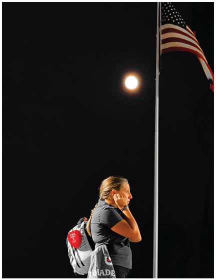 A person with a backpack stands near a flagpole with an American flag at night under a bright full moon. Description generated by AI