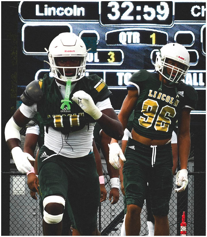 Two people in football uniforms and helmets are walking in front of a scoreboard displaying the team name Lincoln and game details. Description generated by AI