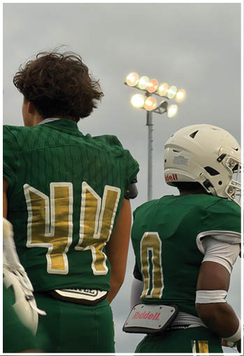 Two people wearing green football uniforms stand side by side under bright stadium lights on a cloudy day. Description generated by AI