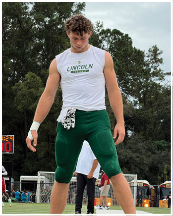 A person wearing a sleeveless white shirt with “LINCOLN" printed on it and green athletic pants stands on a sports field with trees and school buses in the background. Description generated by AI