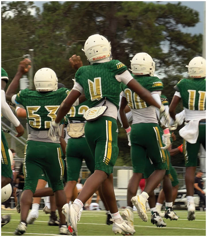 A person wearing a green football uniform with the number 14 is handing a football to another person in a white shirt on empty bleachers under a cloudy sky. Description generated by AI